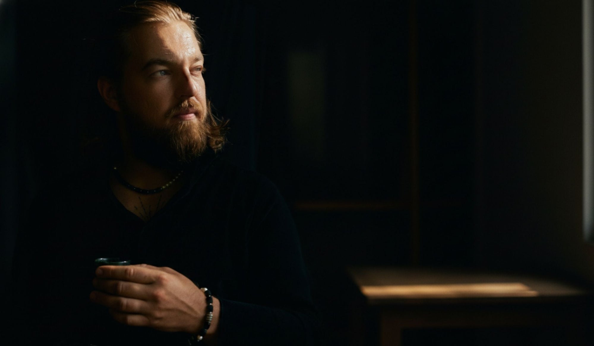 Man sitting in dim light looking away calmly, holding a cup, composed and reflective presence